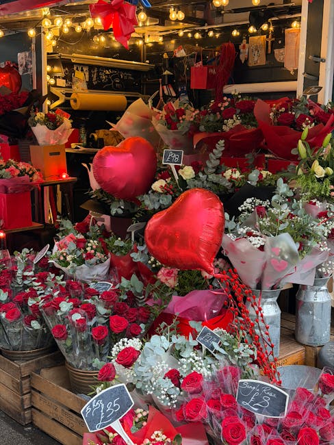 A vibrant display of fresh flowers featuring red roses, white filler flowers, and green foliage arranged in various bouquets and wrapping papers. The arrangement includes heart-shaped red balloons and