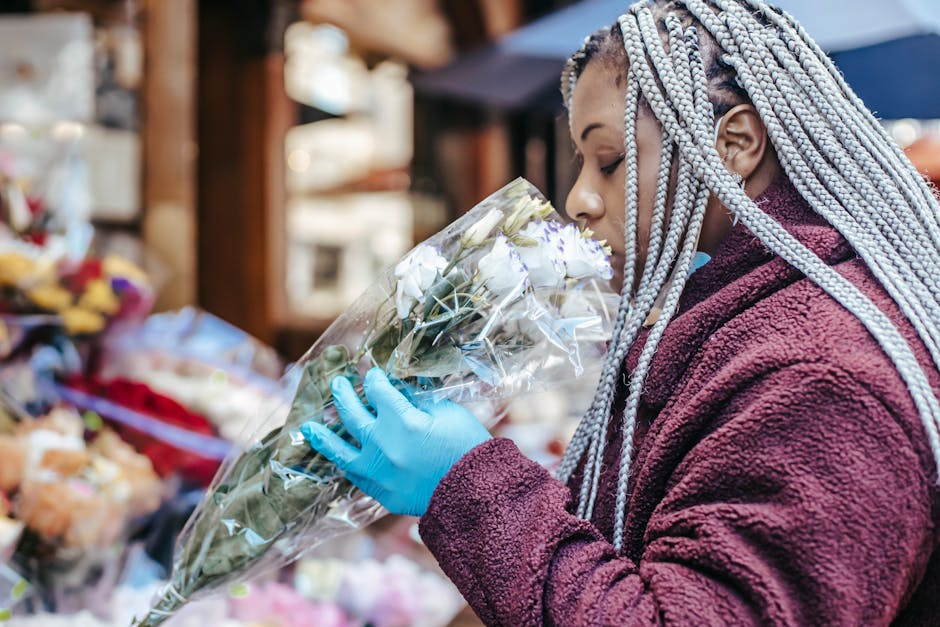 A woman with silver braided hair, wearing a maroon fleece jacket and light blue gloves, is seen holding a bouquet of fresh white roses wrapped in clear plastic at an outdoor flower market. The arrange