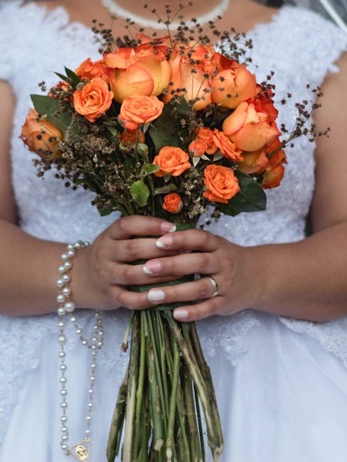 A bride in a white lace wedding dress holds a bouquet of fresh orange roses, peach spray roses, and small sprays of baby's breath, wrapped with a green foliage base. The bouquet is arranged in a round