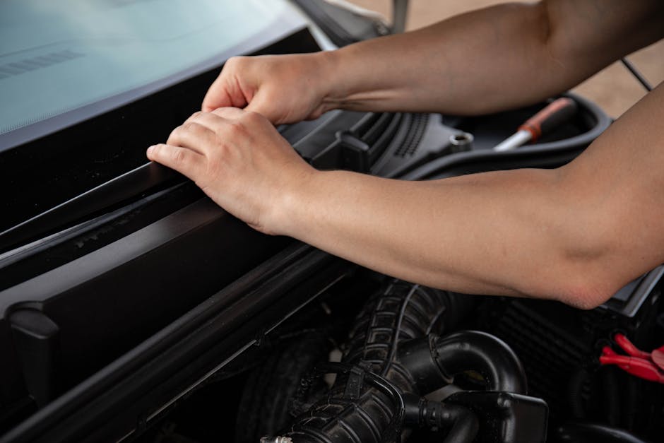 Close-up of a person's hands working on a car engine, with one hand gripping the edge of the engine compartment and the other adjusting or inspecting a component. The image shows a detailed view of th