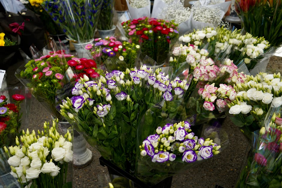A variety of fresh flower bouquets arranged on a display table, featuring purple and white eustoma, white spray roses, pink and white carnations, and small purple daisy-like flowers. The flowers are w