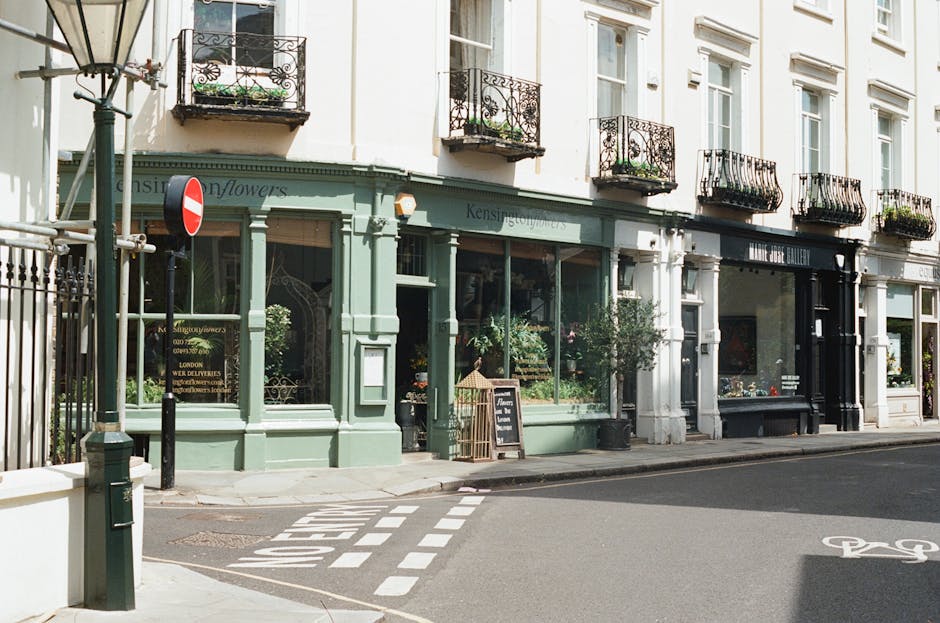 A street view featuring a florist shop with a light green facade and large glass windows displaying floral arrangements inside. The shop is located on the ground floor of a white, multi-story building
