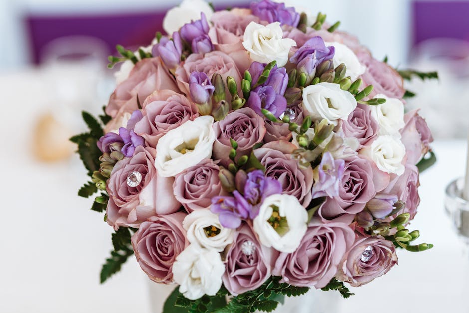 A close-up image of a floral bouquet featuring soft pink roses, white lisianthus, and purple freesias, arranged in a rounded, lush style. The flowers are fresh with vibrant petals and visible green st