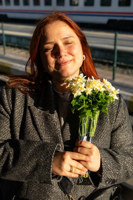 A woman with red hair is holding a fresh bouquet of flowers close to her chest. The bouquet features a mix of white and yellow daffodils, with vibrant green foliage, arranged in a clear glass vase. Sh