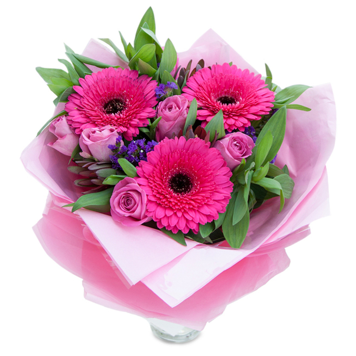 I picture this bouquet as a small, hand-tied embrace: three confident fuchsia Gerbera daisies explode with saturated colour, their radiating petals forming bold, cheerful suns around dark, grounding centres. Surrounding them, a handful of lavender-tinged roses unfold in a softer register - dusty rose and pale mauve tones that curl and catch the light with velvety grace. Interspersed within are compact clusters of purple statice, papery and bright, which create pockets of cool contrast and a whisper of texture against the smoother petals. A generous supporting layer of greenery - broad salal-like leaves and rounded eucalyptus - gives the bouquet structure and a fresh, dewy aroma, suggesting stems just gathered from a local market stall. The flowers sit embraced by an inner sheet of matte pale pink tissue and an outer wrap of translucent fuchsia-pink cellophane that shimmers faintly and reinforces the pink story from bloom to bow. The lower portion reveals the clear glass vase's rim, a practical touch that signals immediacy and display-readiness on a kitchen island or a small dining table. Light in the picture is soft and natural, highlighting petal textures and the gentle folds of the wrapping. Consciously made to lift the spirits, this arrangement would brighten a flat after a stroll along the river near Vauxhall or arrive like a warm note on a damp London morning - ideal for expressing affection, congratulations, or heartfelt thanks with an honest, handcrafted touch.
